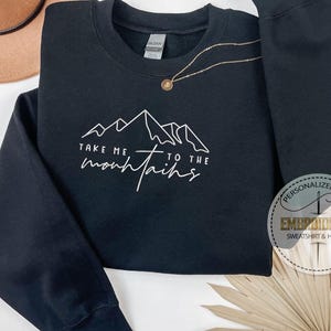 K&ouml;nnte beinhalten: Schwarzes Sweatshirt mit wei&szlig;er Berg-Grafik und dem Text "Take me to the mountains". Eine goldene Halskette mit Anh&auml;nger befindet sich auf dem Sweatshirt. Ein brauner Hut ist im Hintergrund.