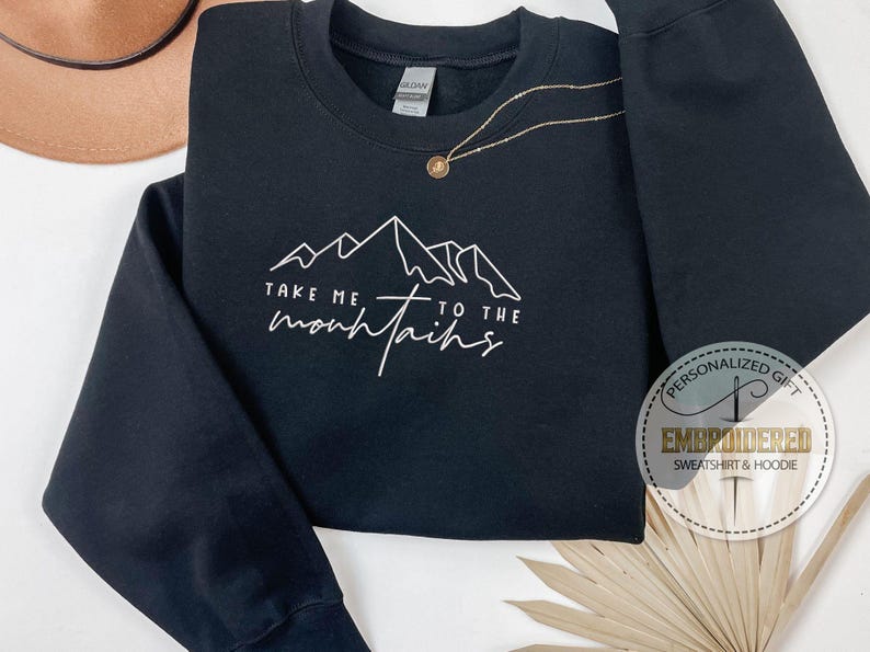 K&ouml;nnte beinhalten: Schwarzes Sweatshirt mit wei&szlig;er Berg-Grafik und dem Text "Take me to the mountains". Eine goldene Halskette mit Anh&auml;nger befindet sich auf dem Sweatshirt. Ein brauner Hut ist im Hintergrund.