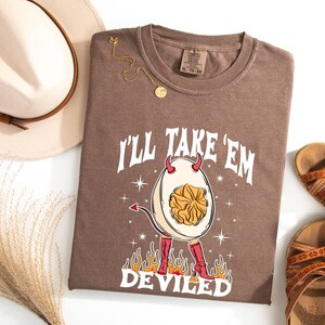 以下が含まれることがあります： 悪魔の角が生えた悪魔の卵の白いグラフィックと「I'll Take 'Em Deviled」というテキストが書かれた茶色のTシャツ。