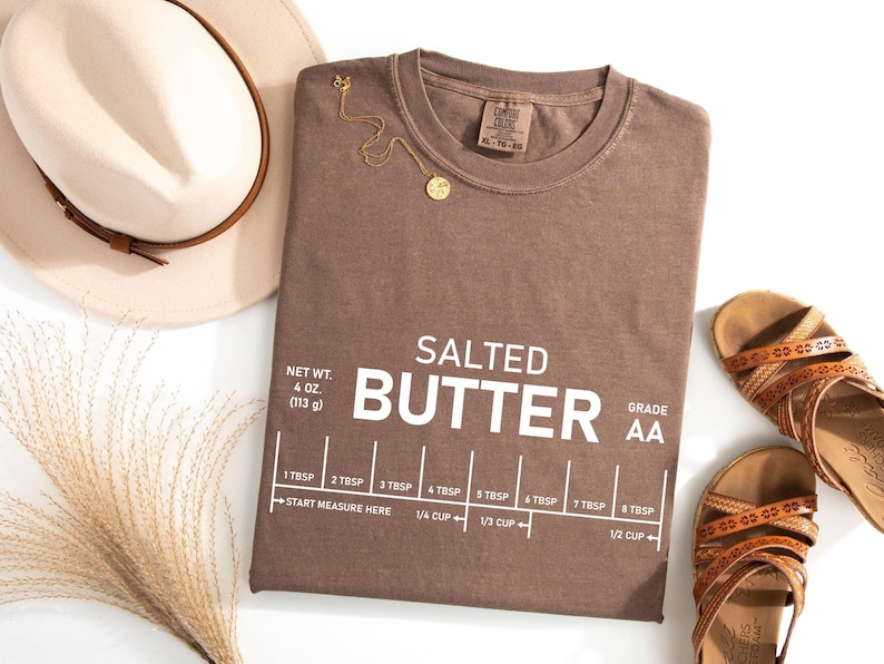 May include: A brown t-shirt with the words "SALTED BUTTER" and a measuring guide printed on it. The shirt also has the text "NET WT. 4 OZ. (113 g)" and "GRADE AA". A gold necklace, a hat, and sandals are also in the image.