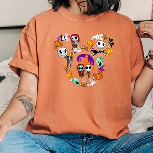 May include: A burnt orange t-shirt with a graphic of Jack Skellington and Sally from The Nightmare Before Christmas in a Mickey Mouse head shape.