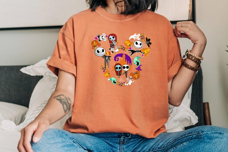 May include: A burnt orange t-shirt with a graphic of Jack Skellington and Sally from The Nightmare Before Christmas in a Mickey Mouse head shape.