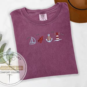 May include: A maroon Comfort Colors t-shirt with embroidered nautical designs: a sailboat, lobster, anchor, and lighthouse. The shirt is folded, with a brown hat in the background. The text "Embroidered Comfort Colors" is in a circle.