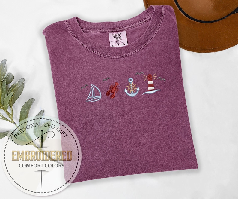 May include: A maroon Comfort Colors t-shirt with embroidered nautical designs: a sailboat, lobster, anchor, and lighthouse. The shirt is folded, with a brown hat in the background. The text "Embroidered Comfort Colors" is in a circle.