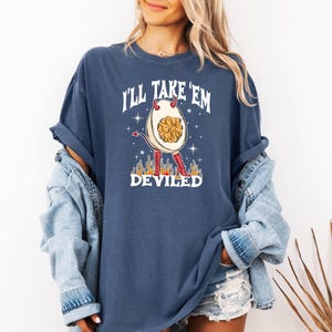 以下が含まれることがあります： 白い悪魔の卵のグラフィックと「I'll Take 'Em Deviled」というテキストが書かれたネイビーブルーのTシャツ。