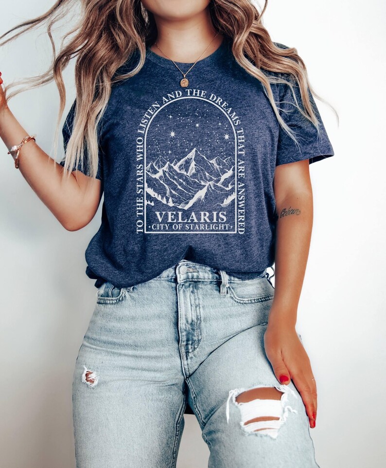 May include: Navy blue t-shirt with a white graphic featuring mountain silhouettes, stars, and the text "TO THE STARS WHO LISTEN AND THE DREAMS THAT ARE ANSWERED". Below the graphic is the text "VELARIS CITY OF STARLIGHT".