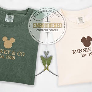 May include: Two folded t-shirts, one olive green and one cream, with embroidered designs. The green shirt features a Mickey Mouse head and the text "MICKEY & CO Est. 1928." The cream shirt has a Minnie Mouse head and "MINNIE & CO Est. 1928."