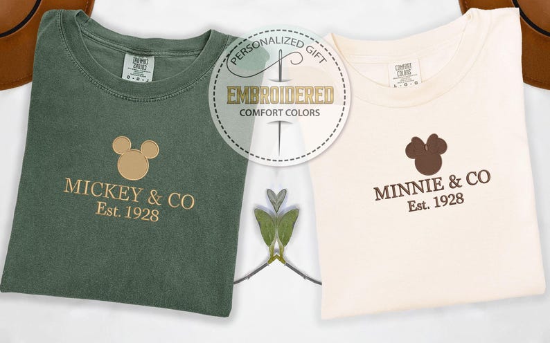 May include: Two folded t-shirts, one olive green and one cream, with embroidered designs. The green shirt features a Mickey Mouse head and the text "MICKEY & CO Est. 1928." The cream shirt has a Minnie Mouse head and "MINNIE & CO Est. 1928."