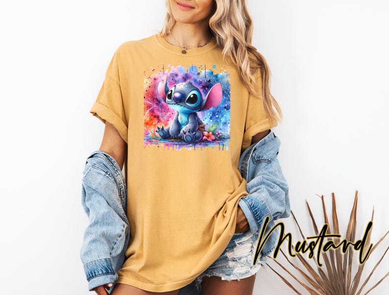 May include: Mustard-coloured t-shirt featuring a watercolour image of Stitch from Lilo & Stitch. The graphic shows Stitch in shades of blue, with a colourful background. The word "Mustard" is written in a script font.