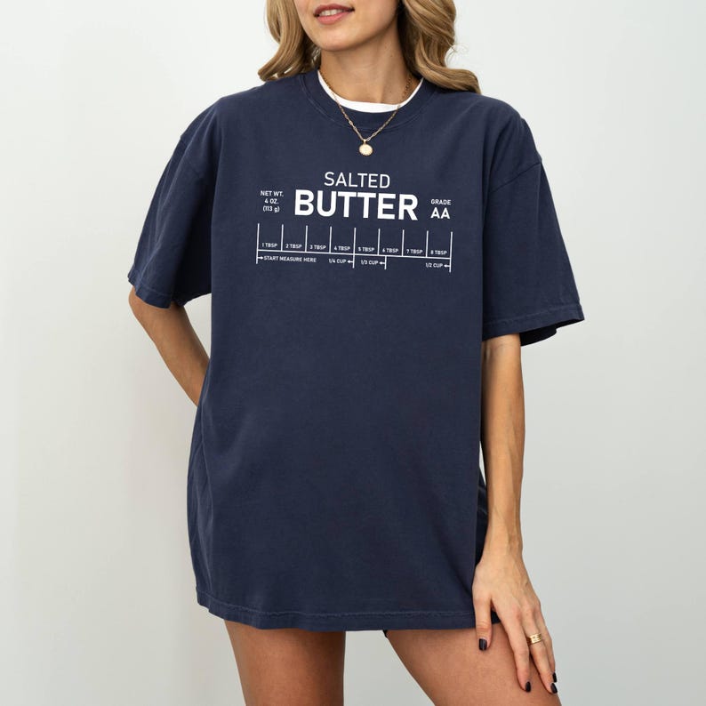 May include: Dark blue t-shirt featuring the text "SALTED BUTTER" and a butter measurement guide in white. The shirt also includes the text "NET WT. 4 OZ (113 g)" and "GRADE AA".