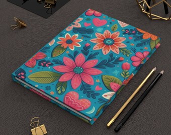 May include: A turquoise blue notebook with a floral pattern featuring pink, orange, and green flowers. The notebook has a white border and is lying on a dark gray surface.