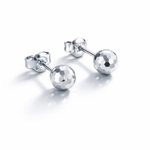 May include: A pair of silver stud earrings. Each earring features a faceted, spherical design. The earrings are displayed on a white background, with the light reflecting off the polished metal. The earrings are small and delicate.