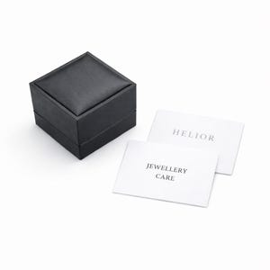 May include: A black jewelry box with a square shape and a padded top. Two white cards with the text "HELIOR" and "JEWELLERY CARE" are next to the box. The box is likely for storing jewelry.