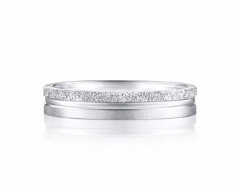 Può includere: Un anello color argento con un design a doppia fascia. La parte superiore presenta una finitura testurizzata e scintillante, mentre la parte inferiore mostra una superficie in metallo opaco e spazzolato. Le due sezioni sono separate da un sottile solco lucido.