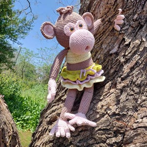 Może przedstawiać: Ręcznie robiona szydełkowa lalka małpki w odcieniach brązu, różu i żółci, umieszczona na pniu drzewa. Małpka ma duże uszy, różowy pysk i nosi żółto-zieloną spódniczkę. To fantazyjny, ręcznie robiony przedmiot.