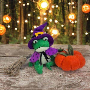 May include: A handmade felt frog wearing a purple witch hat and cape with polka dots, holding an orange pumpkin and a broom. The frog is green and white, with black eyes and a black bow tie. Halloween-themed decor.
