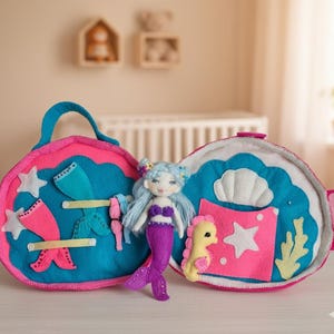 Peut inclure: Un coffret de jeu de sirène en feutre rose et bleu. Le coffret s'ouvre pour révéler une poupée sirène en feutre avec une queue violette, un hippocampe et d'autres accessoires en feutre. Le coffret a une poignée.