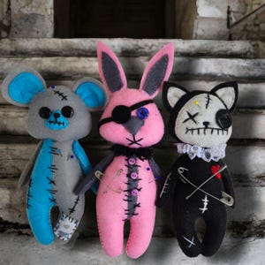May include: Three handmade felt toys: a grey mouse with blue accents, a pink bunny with a black eye patch, and a black cat with a white face and a red heart. Each toy has stitched details and safety pins.