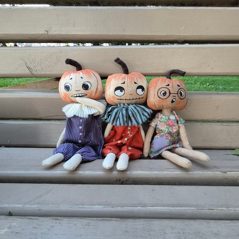 Puede incluir: Tres mu&ntilde;ecas hechas a mano con cabezas de calabaza, caras pintadas y cuerpos de tela sentadas en un banco de madera. Las mu&ntilde;ecas visten diferentes atuendos, incluyendo un mono a rayas moradas, un mono rojo y un vestido floral. Cada mu&ntilde;eca tiene una expresi&oacute;n &uacute;nica.