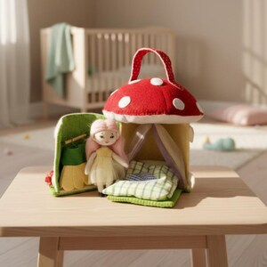 May include: A whimsical toy set featuring a red mushroom house with white polka dots, a doll with pink hair, and a green felt carrying case. The set includes a small bed with a green and white checkered pillow. The toys are displayed on a light wood table.