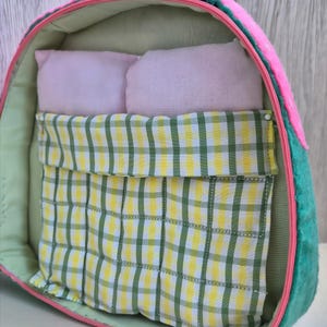 Pu&ograve; includere: Un letto per animali domestici fatto a mano a forma di fetta di anguria. Il letto presenta un esterno rosa, lati verdi e un interno verde chiaro. All'interno ci sono due piccoli cuscini rosa e una coperta a scacchi gialla e verde.