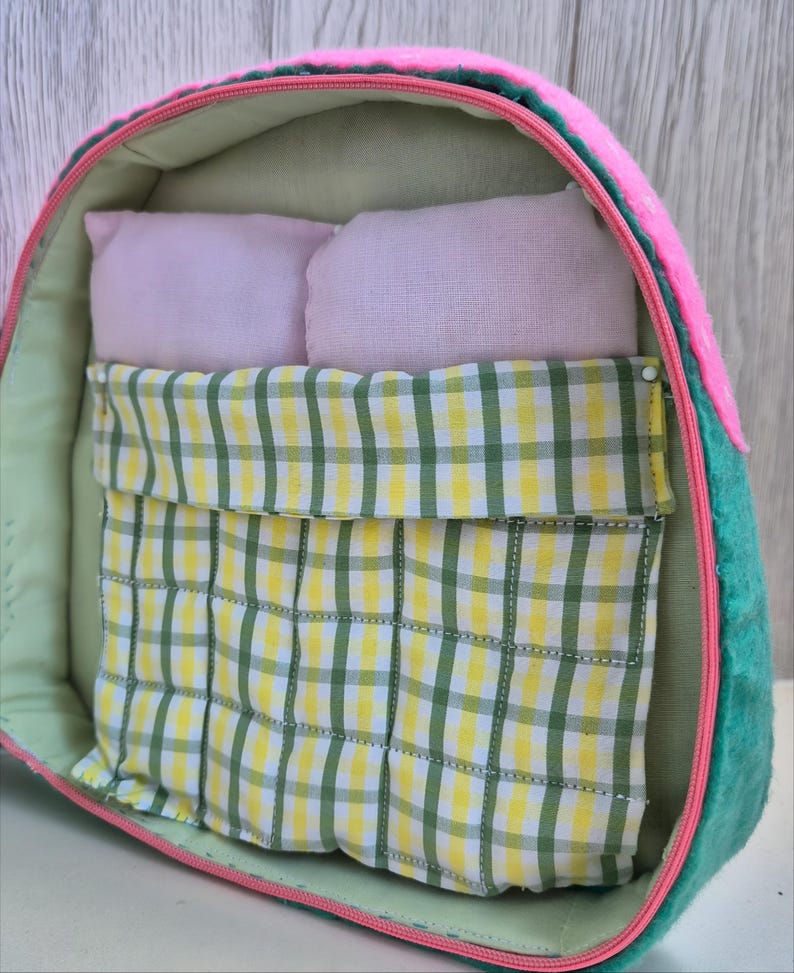 Pu&ograve; includere: Un letto per animali domestici fatto a mano a forma di fetta di anguria. Il letto presenta un esterno rosa, lati verdi e un interno verde chiaro. All'interno ci sono due piccoli cuscini rosa e una coperta a scacchi gialla e verde.