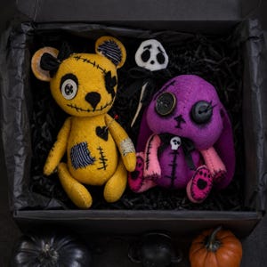 May include: A yellow bear plush with a button eye, stitched mouth, and patches, alongside a purple rabbit plush with button eyes, a skull bow, and stitched details, plus a small white skull ornament, all nestled inside a black box.