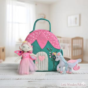 May include: A felt fairy doll with pink wings and a tulle skirt stands beside a felt house-shaped bag with a handle. A felt unicorn with a blue mane and pink accents is also present. The bag is teal and pink with a heart-shaped door.