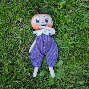 Puede incluir: Una mu&ntilde;eca de peluche hecha a mano con cabeza de calabaza y expresi&oacute;n preocupada. La mu&ntilde;eca viste un mono a rayas moradas y negras con botones coloridos y un cuello blanco con volantes. La mu&ntilde;eca est&aacute; tumbada sobre hierba verde.