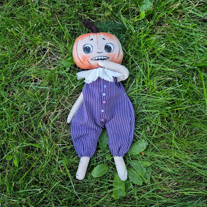 Puede incluir: Una mu&ntilde;eca de peluche hecha a mano con cabeza de calabaza y expresi&oacute;n preocupada. La mu&ntilde;eca viste un mono a rayas moradas y negras con botones coloridos y un cuello blanco con volantes. La mu&ntilde;eca est&aacute; tumbada sobre hierba verde.