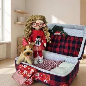 Peut inclure: Une poup&eacute;e faite &agrave; la main avec des cheveux blonds boucl&eacute;s et des lunettes rouges, v&ecirc;tue d'un pull rouge avec un bonhomme de neige, d'un legging rouge et de chaussures blanches. Un chien en peluche avec un chapeau de f&ecirc;te se tient &agrave; c&ocirc;t&eacute; de la poup&eacute;e. La poup&eacute;e est &agrave; c&ocirc;t&eacute; d'une valise ouverte &agrave; carreaux rouges et noirs.