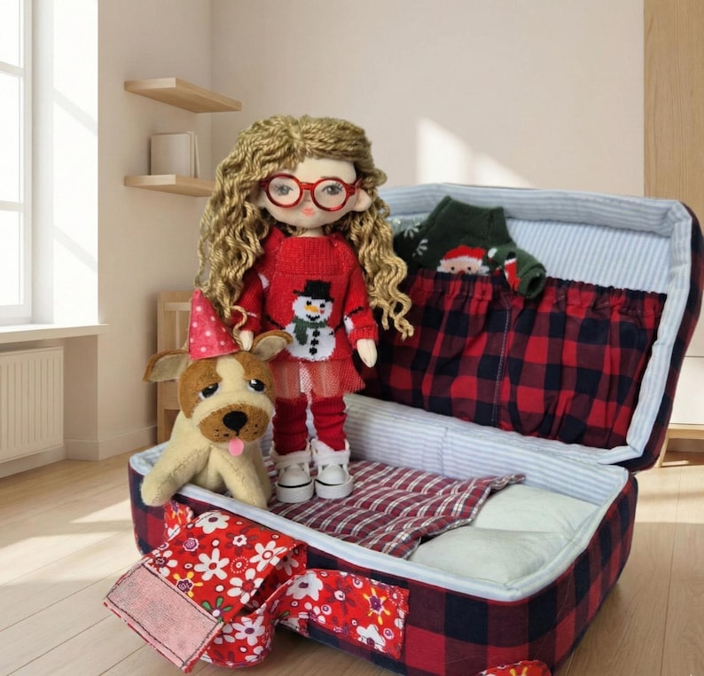 Peut inclure: Une poup&eacute;e faite &agrave; la main avec des cheveux blonds boucl&eacute;s et des lunettes rouges, v&ecirc;tue d'un pull rouge avec un bonhomme de neige, d'un legging rouge et de chaussures blanches. Un chien en peluche avec un chapeau de f&ecirc;te se tient &agrave; c&ocirc;t&eacute; de la poup&eacute;e. La poup&eacute;e est &agrave; c&ocirc;t&eacute; d'une valise ouverte &agrave; carreaux rouges et noirs.