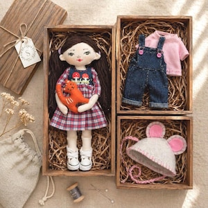 Peut inclure: Une poupée artisanale aux cheveux bruns et une robe à carreaux, tenant un jouet chat orange, est présentée dans une boîte en bois. D'autres boîtes contiennent une salopette en jean, un haut rose et un chapeau à oreilles de souris. Un coffret cadeau avec une étiquette est également inclus.