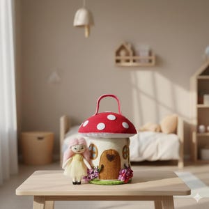 May include: A whimsical felt mushroom house with a red cap and white polka dots, a brown door, and a handle. A small doll with pink hair and a yellow dress stands beside the house. The house is decorated with flowers.