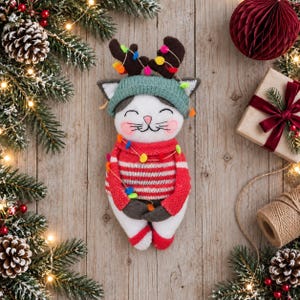 May include: A plush cat toy wearing a teal knit beanie with brown reindeer antlers, a red and white striped sweater, and red-tipped paws. The cat is decorated with a string of colorful miniature pom-pom lights draped over its head and body.
