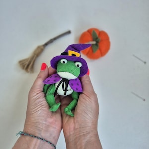 May include: A handmade green frog plushie wearing a purple witch hat and cape with polka dots. A small orange pumpkin and a miniature broom are in the background. The frog has large, round eyes and a black bow tie.