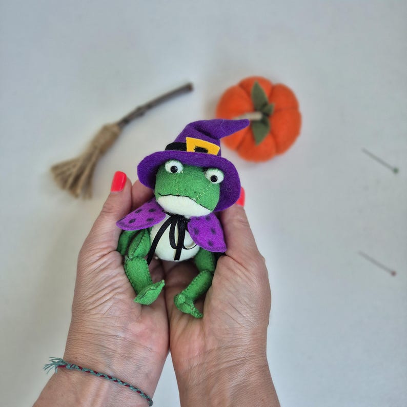 May include: A handmade green frog plushie wearing a purple witch hat and cape with polka dots. A small orange pumpkin and a miniature broom are in the background. The frog has large, round eyes and a black bow tie.