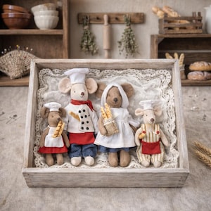May include: Four plush mice dressed as bakers, chefs, and servers, holding miniature baguettes. They are arranged in a wooden tray with a lace doily, set against a rustic kitchen backdrop with shelves and bread.