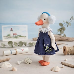 Pode incluir: Boneca de pato feita à mão, vestindo um vestido azul marinho com bordados florais brancos, uma gola de renda branca e uma faixa azul com protetores de ouvido. O pato tem bico e pés laranja. O fundo inclui uma pintura de farol e decoração náutica.