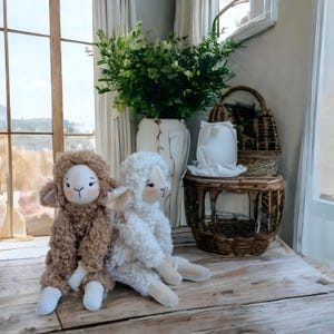 May include: Two plush sheep dolls, one brown and one white, sit side-by-side on a wooden surface. The dolls have soft, textured fur, pink cheeks, and embroidered facial features. The background includes a vase with greenery and wicker baskets.