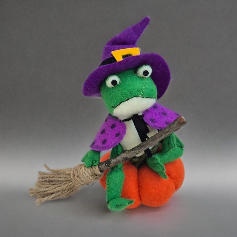 May include: A whimsical Halloween decoration featuring a green frog dressed as a witch. The frog wears a purple witch hat with a yellow buckle, a purple cape with polka dots, and sits on an orange pumpkin, holding a broom.