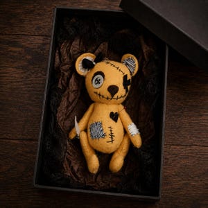 Könnte beinhalten: Ein gelber Teddybär aus Filz mit einem Knopfauge, einem aufgestickten schwarzen X-Auge und einem aufgestickten Mund. Der Bär hat eine schwarze Schleife, einen Herzflicken und gestreifte Stoffflicken an Körper und Ohr, präsentiert in einer schwarzen Box.