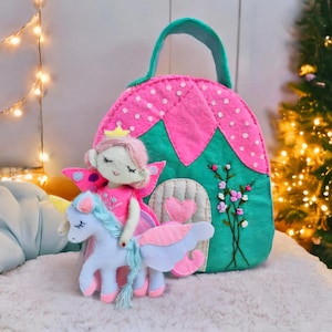 Pu&ograve; includere: Una bambola fata in feltro fatta a mano con un unicorno e una borsa a forma di casa rosa e verde. La fata ha ali rosa e una corona d'oro. La borsa ha un manico e una porta con un cuore. L'unicorno &egrave; azzurro con accenti rosa.