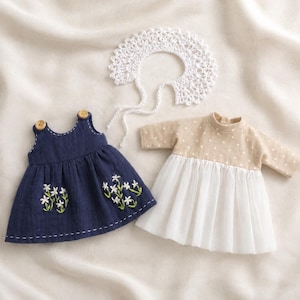 Puede incluir: Un vestido azul oscuro sin mangas con bordado floral blanco y dos botones de madera, un vestido beige con lunares blancos y falda blanca, y un cuello de ganchillo blanco con lazos largos, todo diseñado para una muñeca o figura pequeña.