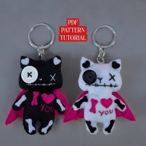 May include: Two felt cat keychains with pink capes and black and white bone patterns. The black cat has a white button eye and an X for the other eye. The white cat has a black button eye and an X for the other eye. Both cats have a pink heart with the text "I love you" on their chests.