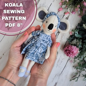 May include: A white koala doll wearing a blue and white patterned dress and a blue headband. The doll is 8 inches tall and is being held by a person's hands.