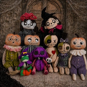May include: A collection of handmade fabric dolls featuring various characters including pumpkin-headed figures in ruffled collars, a green frog with a broom, a purple rabbit, and stitched rag dolls with button eyes, all displayed together.