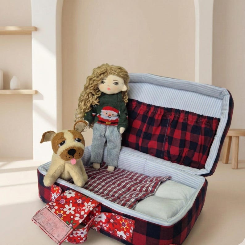 Peut inclure: Une poup&eacute;e aux cheveux blonds boucl&eacute;s, v&ecirc;tue d'un pull vert avec un motif de P&egrave;re No&euml;l et d'un pantalon gris, se tient dans une valise ouverte. Un chien en peluche et une sangle &agrave; motifs floraux rouges et blancs sont &eacute;galement dans l'image. La valise a un motif &agrave; carreaux rouges et noirs.
