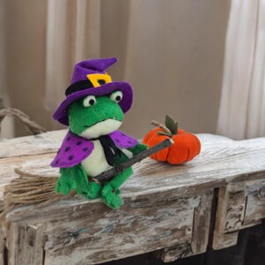 May include: A handmade green frog wearing a purple witch hat and cape with a black tie, holding a wooden broom. An orange pumpkin with a green stem sits nearby. The frog is a Halloween decoration.