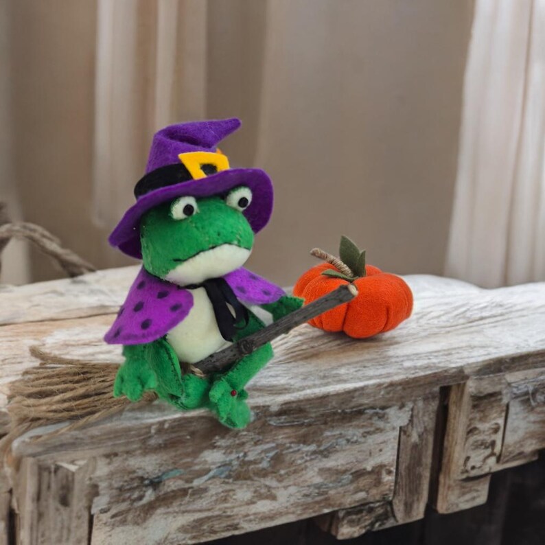 May include: A handmade green frog wearing a purple witch hat and cape with a black tie, holding a wooden broom. An orange pumpkin with a green stem sits nearby. The frog is a Halloween decoration.